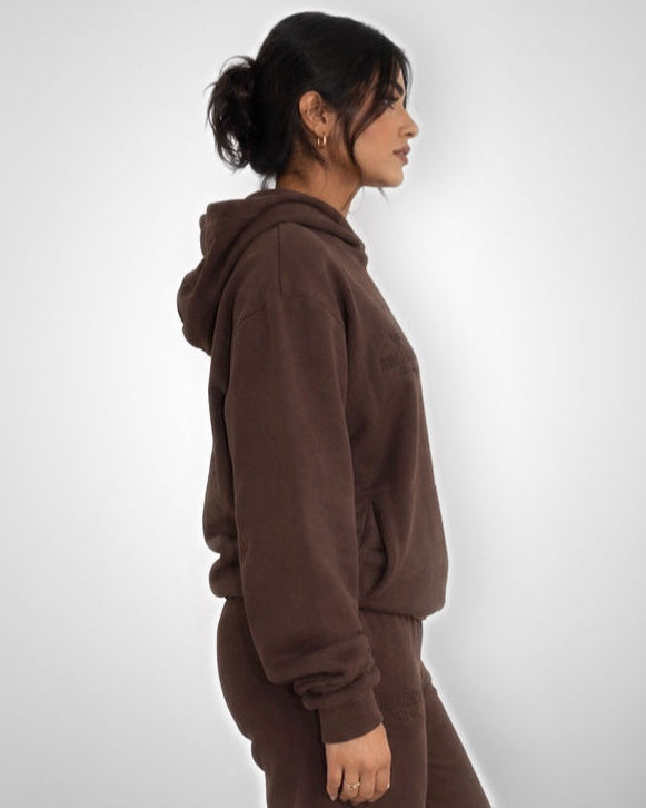 A woman shown in side profile wearing a matching brown hooded sweatshirt and jogger pants against a light gray studio background.