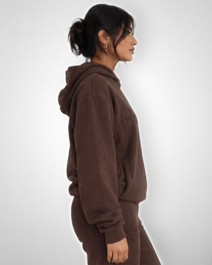 A woman shown in side profile wearing a matching brown hooded sweatshirt and jogger pants against a light gray studio background.