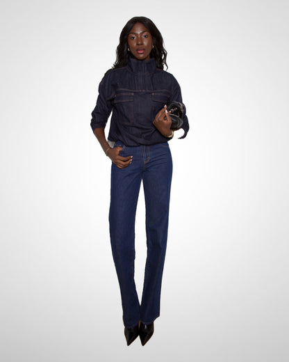A model stands against a light gray studio background wearing a dark indigo denim jacket and matching dark blue straight-leg jeans, with black pointed shoes and a small black clutch.