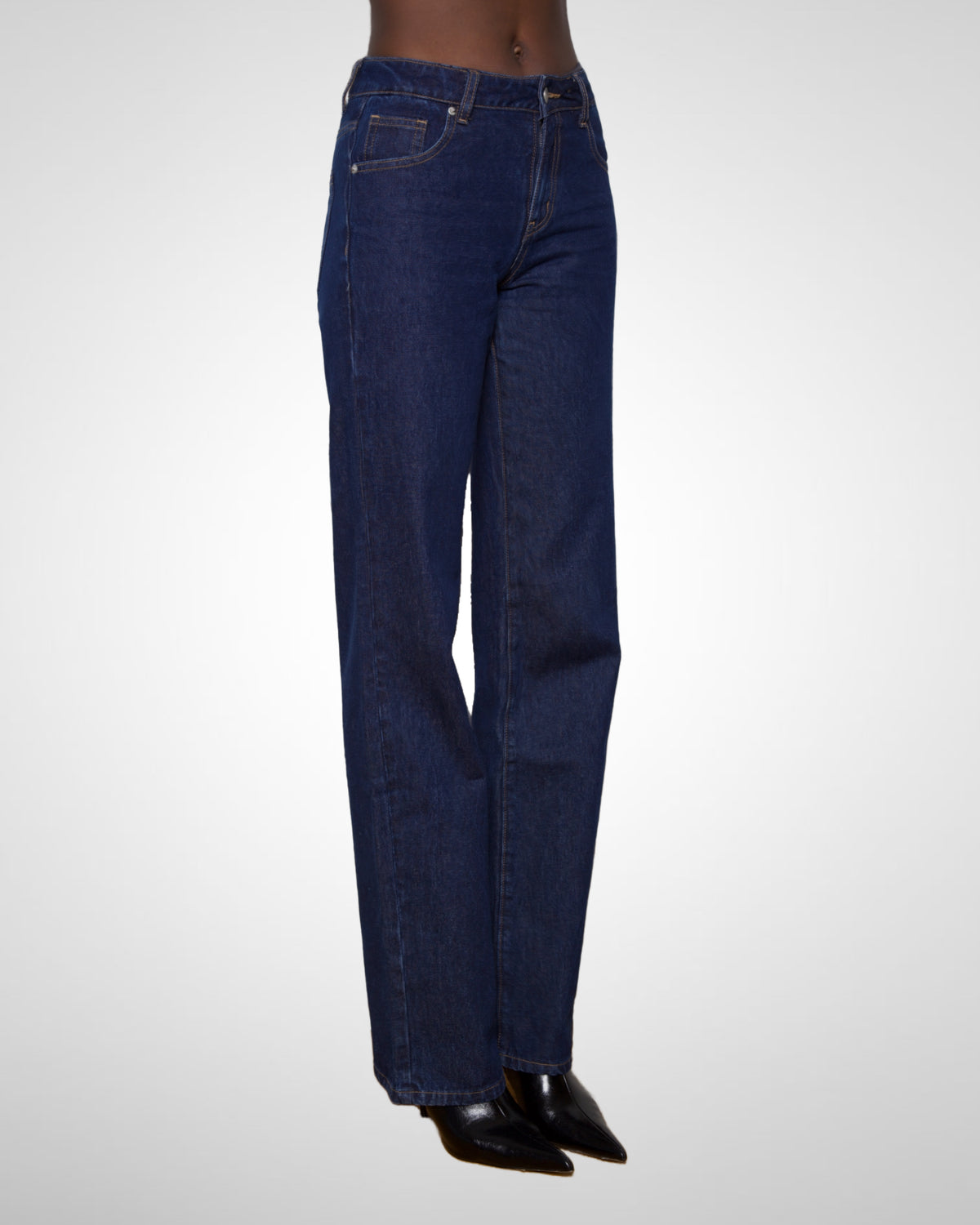 A tall woman  wearing dark blue straight-leg denim jeans and black pointed shoes, shown from the waist down against a light gray studio background.