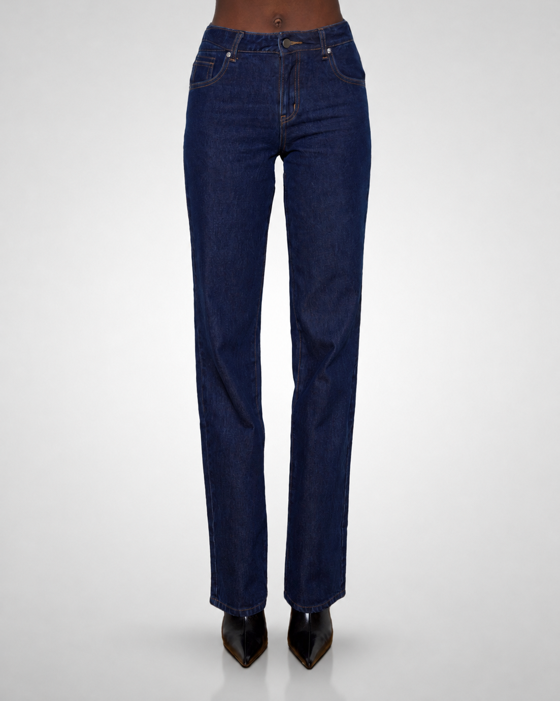 A tall model is shown from the waist down wearing dark blue straight-leg denim jeans and black pointed-toe heels, photographed against a light gray studio background.