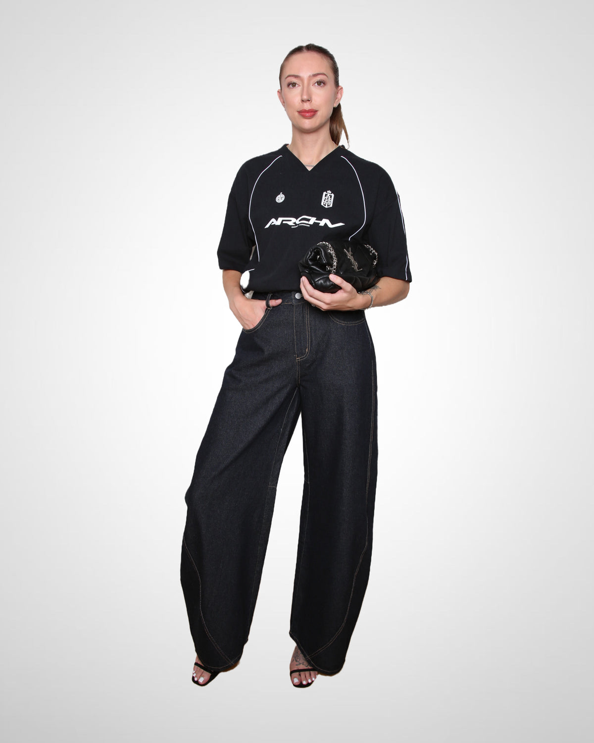 A model stands against a light gray studio background wearing a black short-sleeve top with white piping and the brand name 'ARCHN' across the chest, paired with wide-leg dark jeans and holding a black textured clutch.
