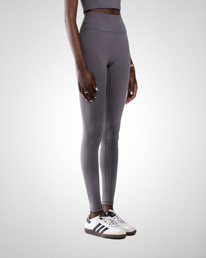 A model wearing tall high-waisted, form-fitting leggings in a dark gray color, paired with white sneakers, shown from the waist down against a light gray studio background.