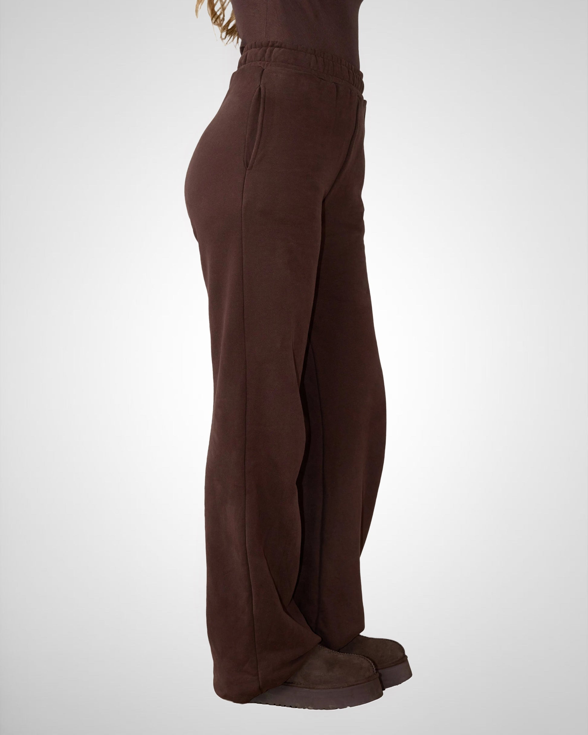 A side profile view of a person wearing dark brown wide-leg lounge pants and matching brown slippers against a light gray studio background.