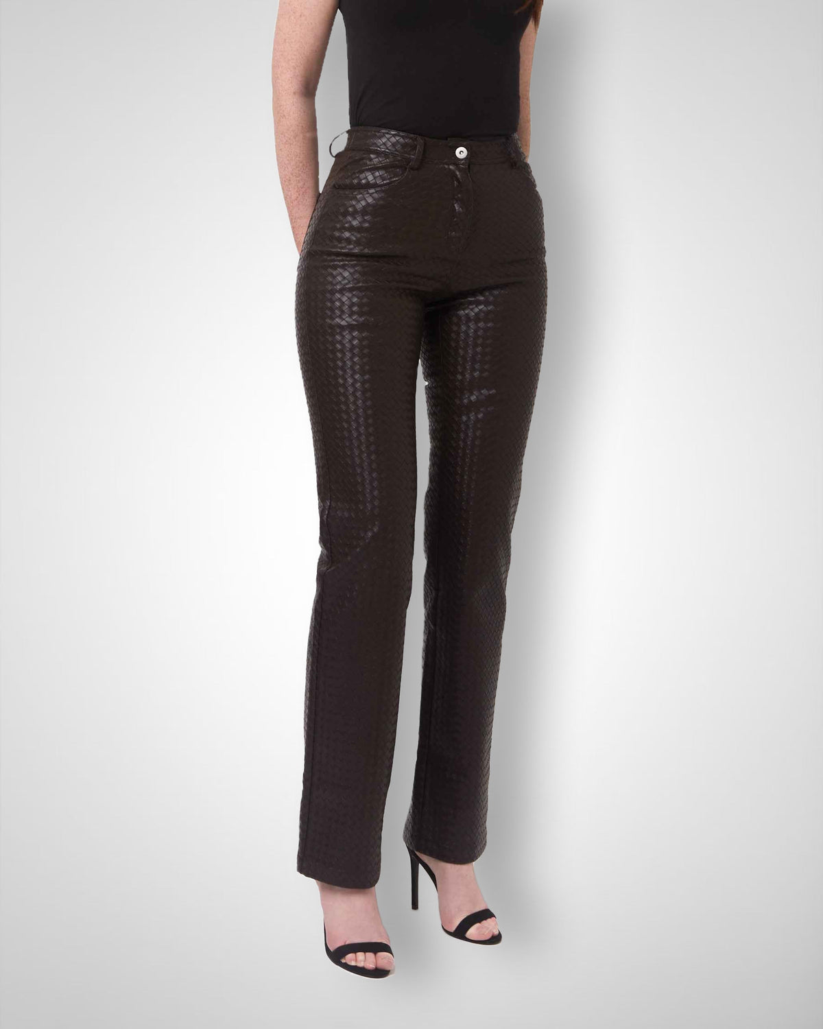A model wearing dark brown textured leather pants with a subtle basketweave pattern, paired with a black top and black high-heeled sandals, shown against a light gray studio background.